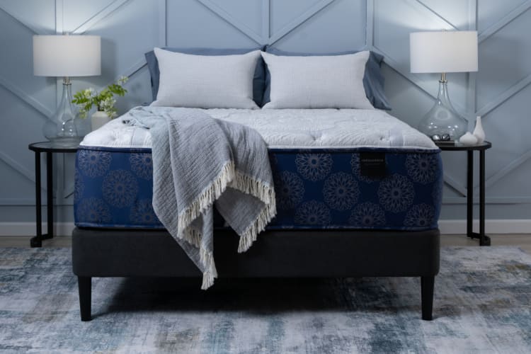 King-size Indulgence mattress with blue floral pattern and plush white top, set on a black upholstered bed frame with modern side tables and lamps.