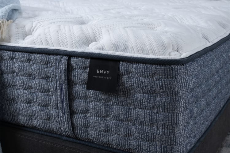 Close-up of an Envy brand mattress corner featuring a textured blue fabric side panel and a white quilted top with the label "ENVY WELCOME TO BED."