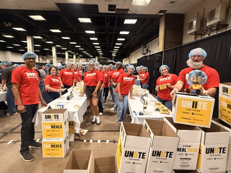 A group of volunteers in red Bob's Community Outreach T-shirts and blue hairnets packing meals at a large indoor event with stacked boxes labeled "UNITE FOR GOOD 20M MEALS" by 9.11 Day, AmeriCorps, Reliance, and Delta.