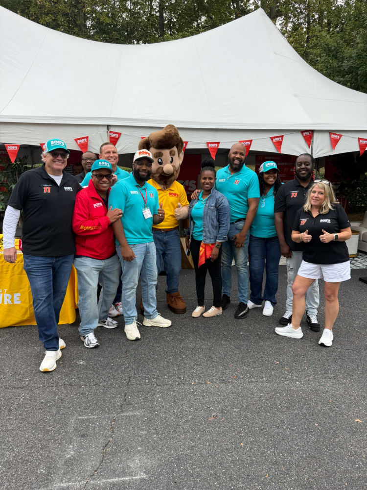 Group of diverse people posing outdoors under a white event tent, wearing branded apparel and hats with "BOB'S" logos, promoting a community or company event.