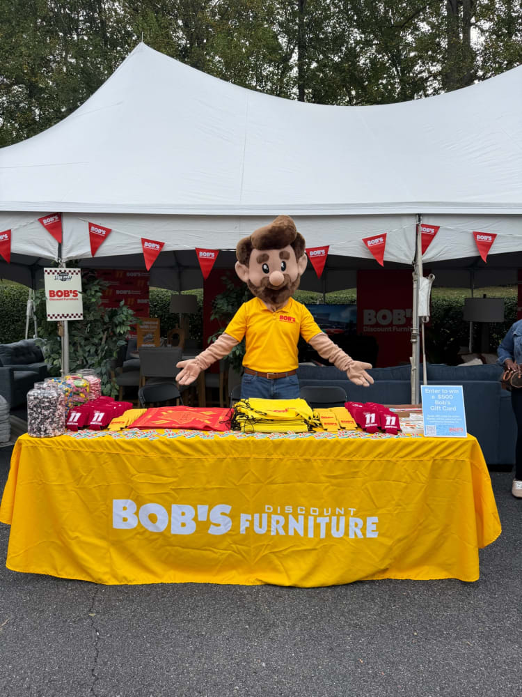 Mascot wearing a yellow Bob's Discount Furniture shirt stands behind a yellow tablecloth-covered table with Bob's Discount Furniture logo, promotional items, and a sign to enter a $500 gift card contest under a white event tent.