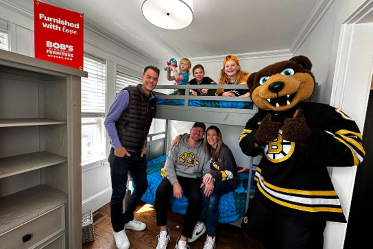 A furnished bedroom with a gray bunk bed and blue bedding, featuring a group of people and a mascot in a Boston Bruins hockey jersey. A red sign reads "Furnished with love - Bob's Discount Furniture "