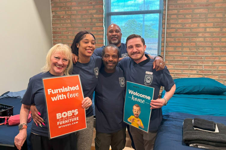 Group of five people wearing navy blue shirts holding signs with texts "Furnished with love Bob's Discount Furniture Mybobs.com" and "Welcome home" in a room with brick wall and blue bedding.