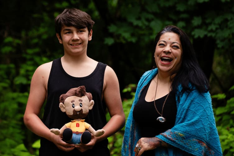 Two people stand outdoors against a green foliage background; one holds a small plush doll with brown hair, beard, yellow shirt, and blue pants, both wearing casual clothing.