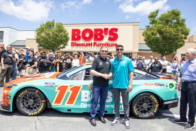 Two men shaking hands in front of a turquoise and orange NASCAR race car with number 11, parked outside a Bob's Discount Furniture store.