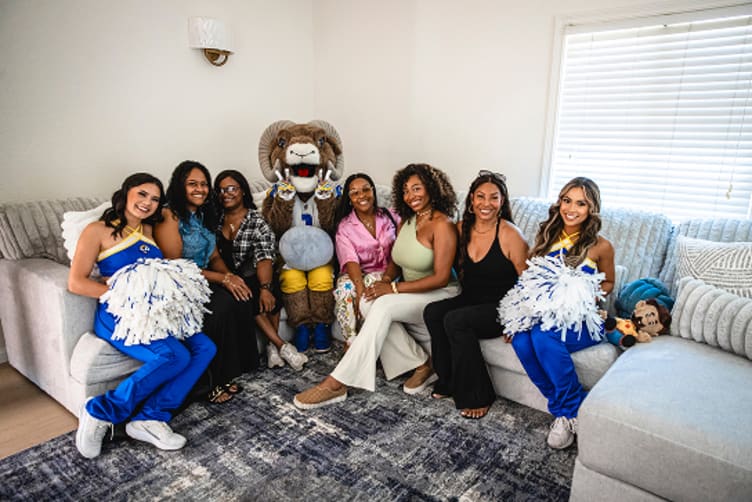 A group of seven women and a person in a ram mascot costume sitting on a gray sectional sofa in a bright living room, two women in blue and yellow cheerleading uniforms holding white pom-poms.