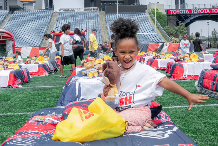 A child wearing a white t-shirt sits on a Patriots-themed inflatable bed on a football field, holding a plush toy. Multiple similar beds with plush toys and yellow bags are arranged on the field, indicating a community or charity event at a stadium.