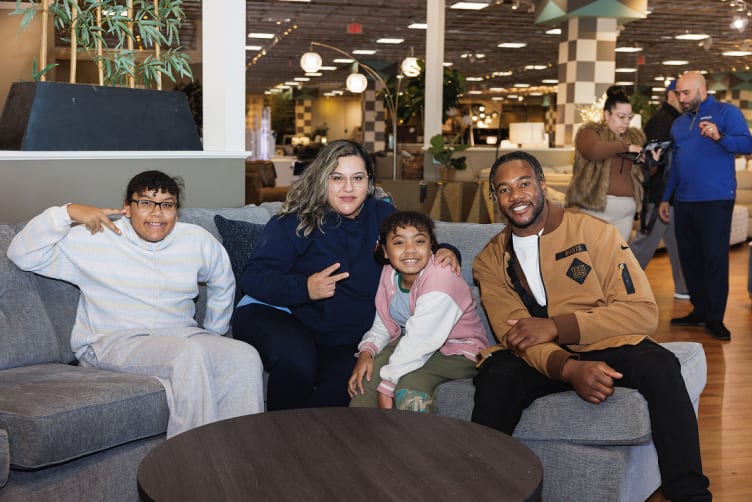 A diverse group of five people sitting on a gray sectional sofa in a furniture store showroom, posing casually with hand gestures.