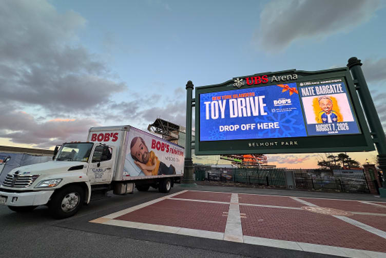 UBS Arena digital billboard at sunset promoting New York Islanders Toy Drive with drop-off details alongside a Bob's Moving truck.