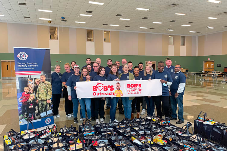 Group of people wearing matching navy blue shirts holding a banner that reads "BOB'S Outreach" and "Bob's Discount Furniture Mybobs.com" with bags of groceries in front, promoting support for military families through Operation Homefront.
