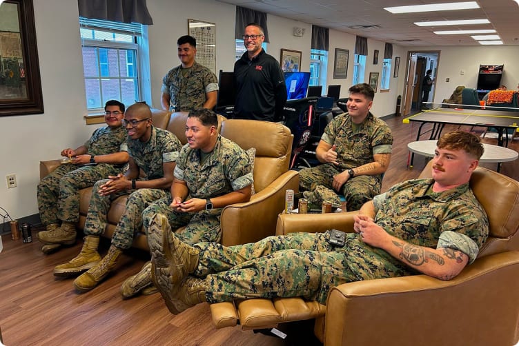 Group of six military personnel in camouflage uniforms and boots relaxing in a lounge with leather chairs, wooden floor, and a ping pong table in the background.