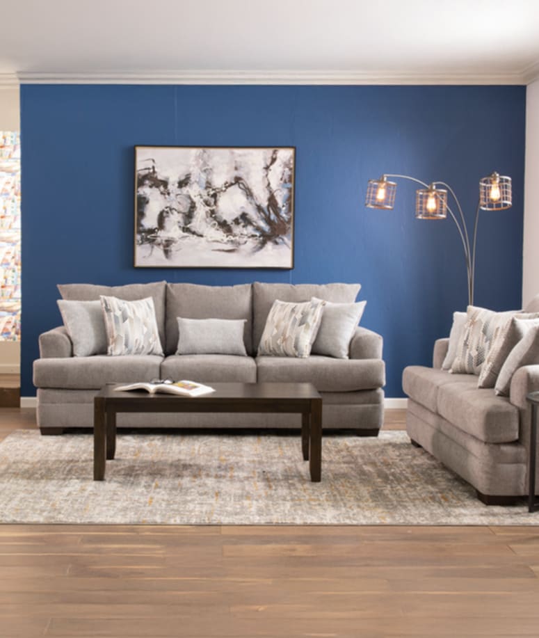 A cozy living room features a gray sofa set with patterned pillows, a dark wood coffee table, and a modern floor lamp against a blue accent wall and abstract art.