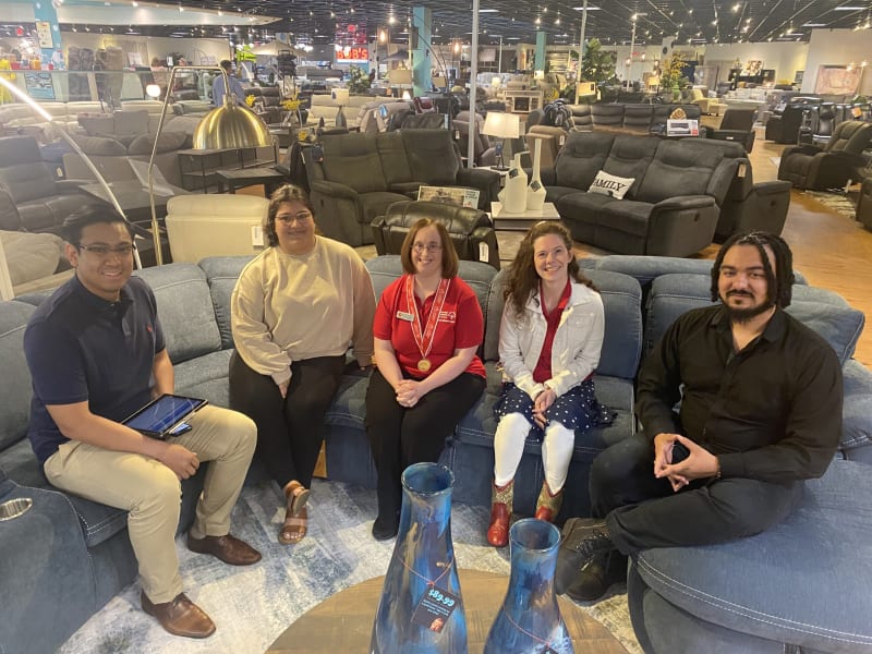 Five people seated on a large blue sectional sofa in a furniture store showroom, surrounded by various couches, lamps, and home decor items.
