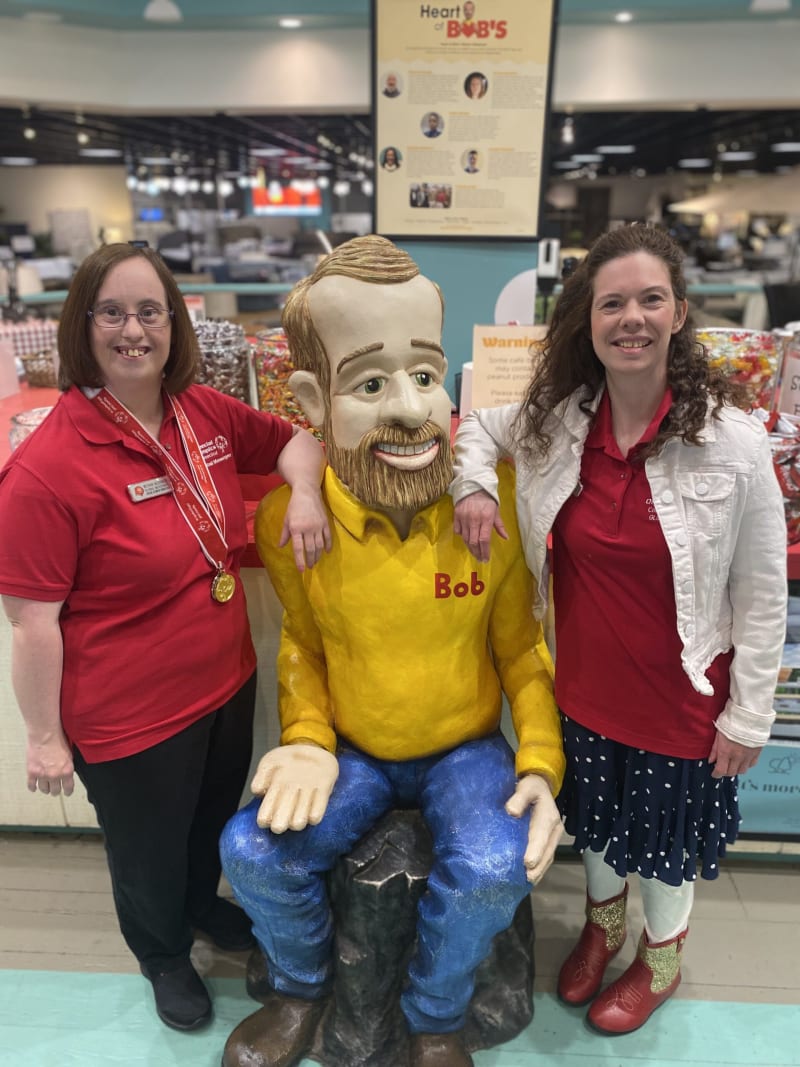 Two women in red polo shirts stand on either side of a seated Bob's Big Boy statue wearing a yellow shirt with "Bob" embroidered, blue pants, and brown shoes, inside a candy store with jars of colorful candy in the background.