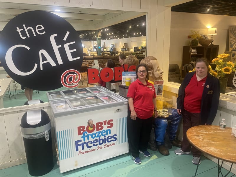 Two employees in red shirts stand beside a white freezer labeled "BOB'S frozen Freebies Premium Ice Cream" under a large black sign reading "the Café @ BOB'S" in a café setting.
