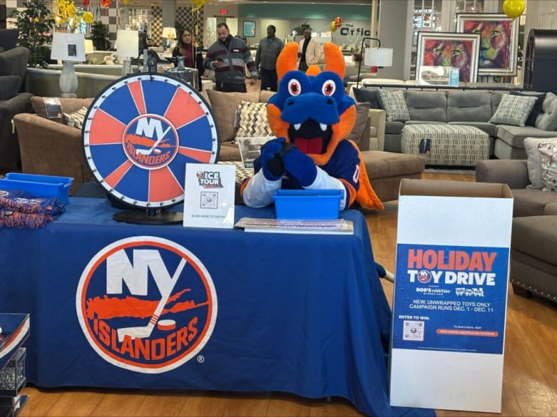 NY Islanders holiday toy drive setup with mascot in blue and orange costume, prize wheel, and donation box reading: "HOLIDAY TOY DRIVE. NEW, UNWRAPPED TOYS ONLY. CAMPAIGN RUNS DEC. 1 - DEC. 11."
