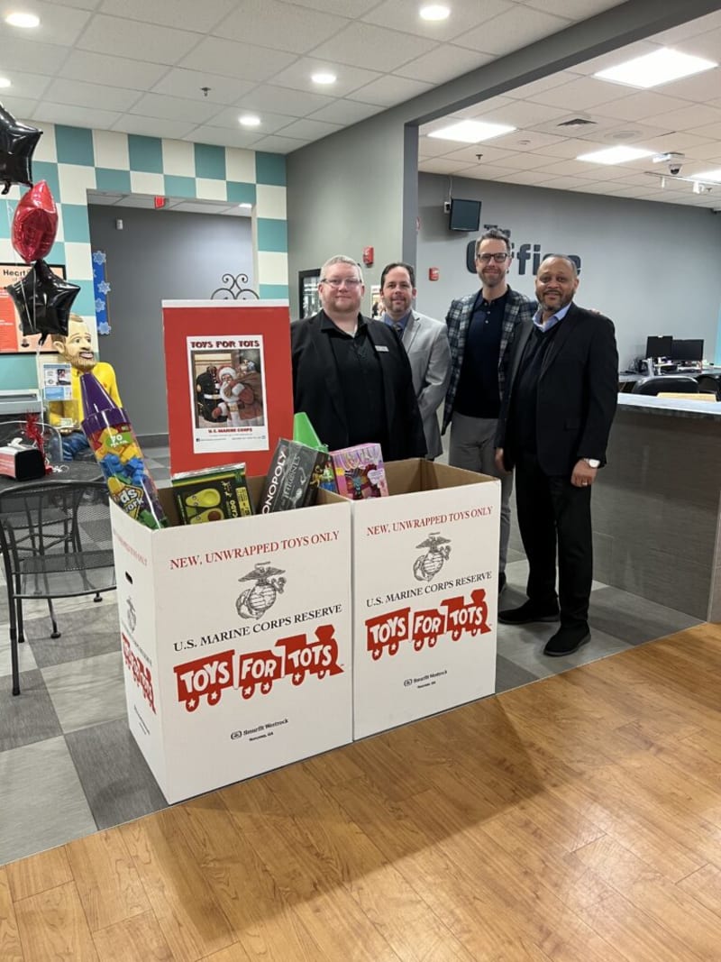 Four men in business attire stand behind two large white U.S. Marine Corps Reserve Toys for Tots donation boxes filled with toys, in an indoor office setting.