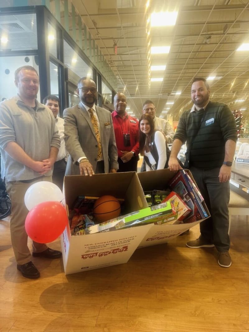 Group of seven people standing indoors around a large Toys for Tots donation box filled with toys including a basketball, board games, and balloons, in a well-lit retail or event space.