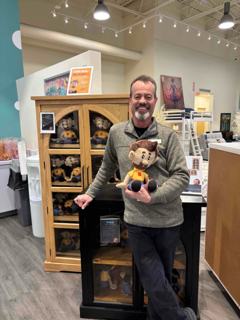 Man wearing gray fleece sweater and black pants holding a plush doll with brown hair, mustache, and yellow shirt with red "B" logo, standing in front of wooden and black display cabinets filled with identical plush dolls in a retail store. Sign on top of cabinet reads "Little Bob, Big Impact."