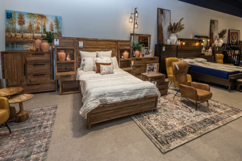 Modern bedroom furniture set in a showroom featuring wooden bed frames, matching nightstands, dressers, and armchairs on patterned rugs.