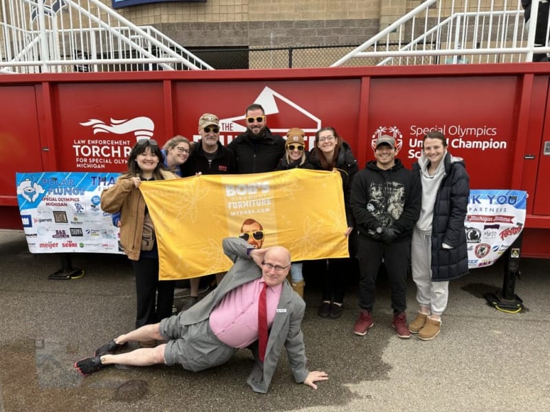 Group of people posing in front of a red law enforcement torch run vehicle for Special Olympics Michigan, holding a yellow Bob's Discount Furniture banner.