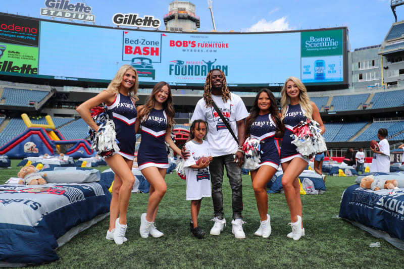 Cheerleaders in navy blue Patriots uniforms with pom-poms pose on Gillette Stadium field with a man and child in Bed-Zone Bash event shirts.