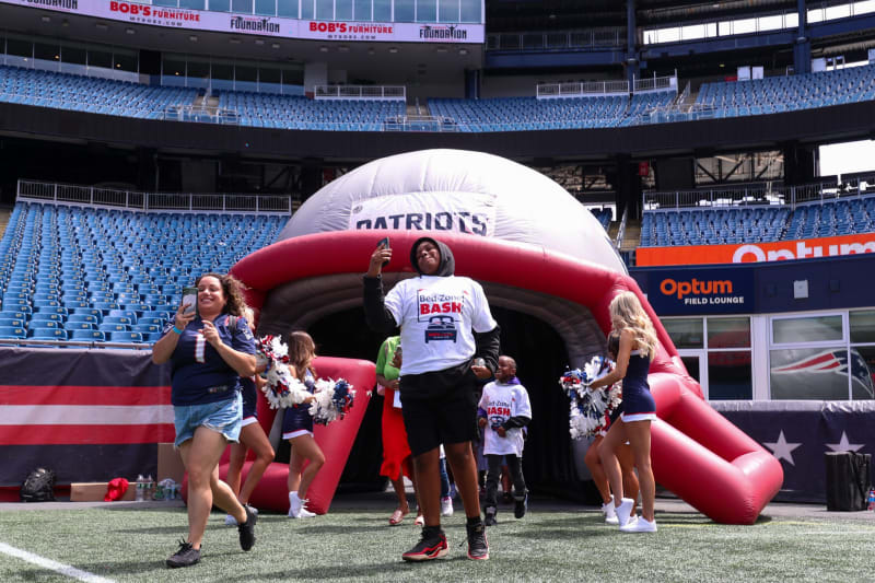 People at a football stadium near a large inflatable Patriots helmet tunnel with "Bed-Zone BASH" shirts, cheerleaders, and fans.