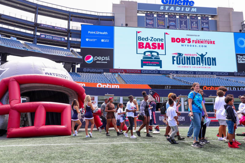 Crowd of people walking on the field at Gillette Stadium near a large inflatable Patriots helmet and a digital billboard displaying "Bed-Zone Bash," "Bob's Discount Furniture," and "Patriots Foundation" logos and text.