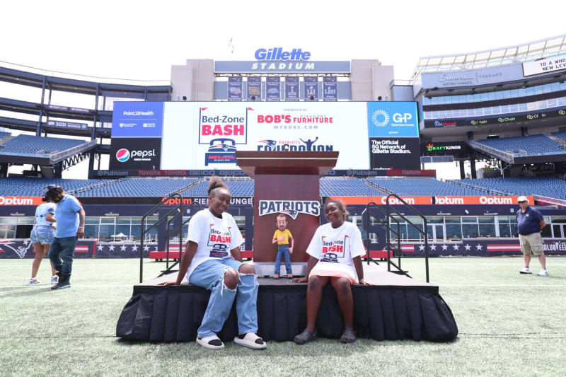 Two children wearing white "Bed-Zone Bash" t-shirts sit on a stage in front of a podium with the Patriots logo at Gillette Stadium. The large stadium screen behind them displays "Bed-Zone Bash," "Bobs Furniture," and "GP" logos, promoting a Patriots Foundation event.