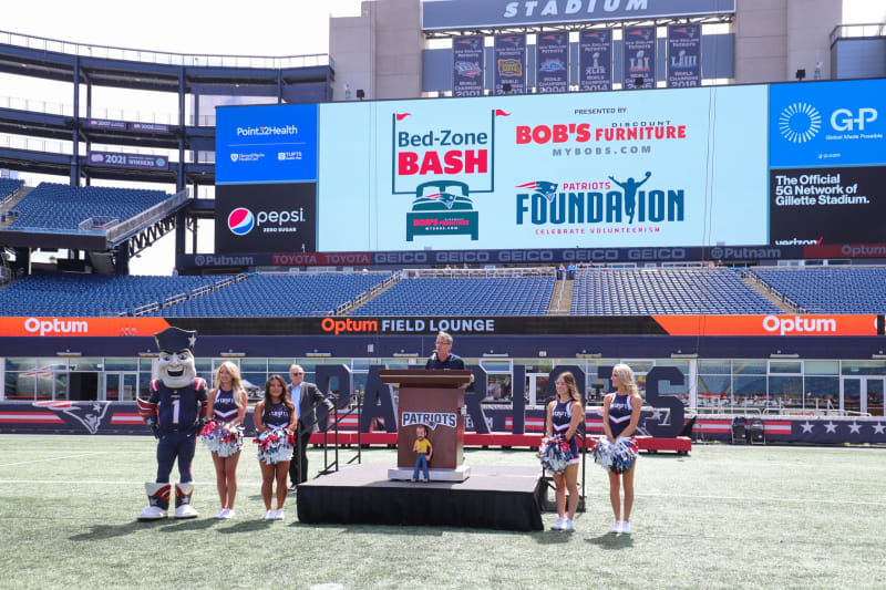 Patriots mascot and cheerleaders stand on Gillette Stadium field near podium with "Patriots" logo, large screen shows Bed-Zone Bash, Bob's Furniture, Patriots Foundation logos.