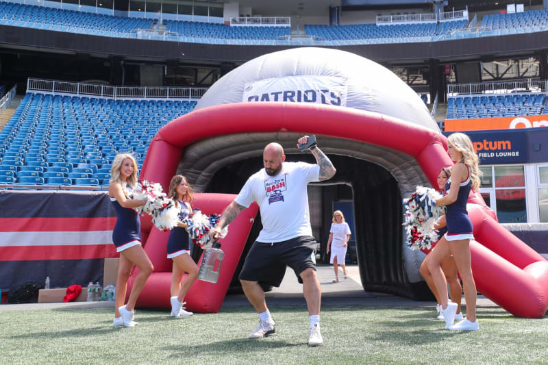 Man in white "Beer Zone Bash" t-shirt and black shorts posing with cheerleaders holding red, white, and blue pom-poms in a stadium near a red and white inflatable Patriots helmet tunnel.