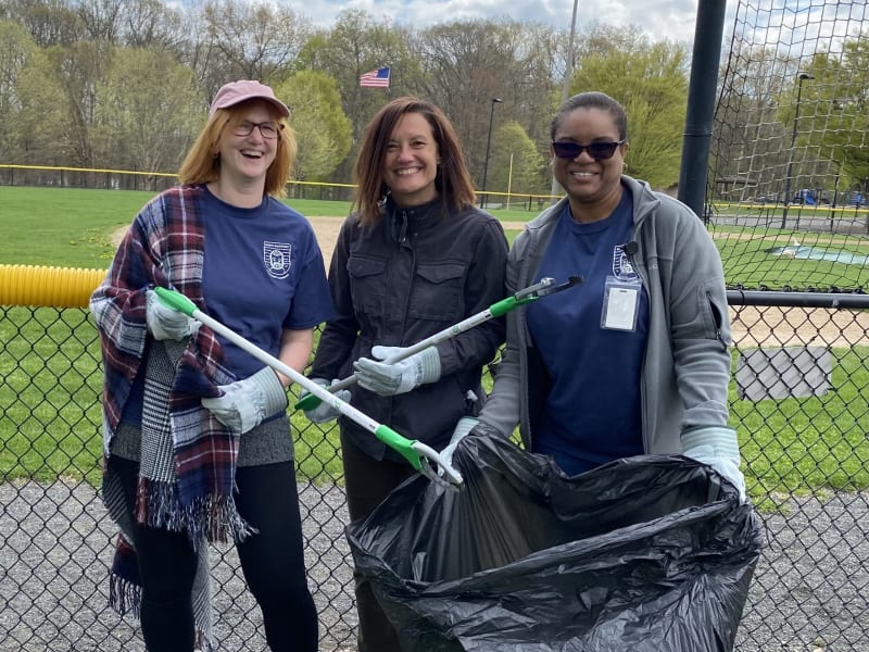 Three people wearing gloves and holding litter pickers and a large black trash bag, participating in outdoor park cleanup on a cloudy day.