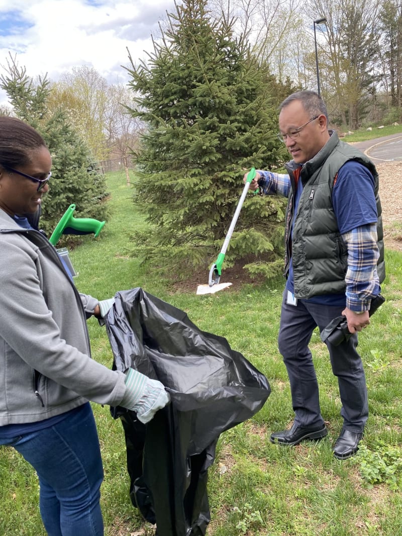Two people outdoors collecting litter with a black trash bag and a green-handled litter picker, surrounded by grass and evergreen trees.