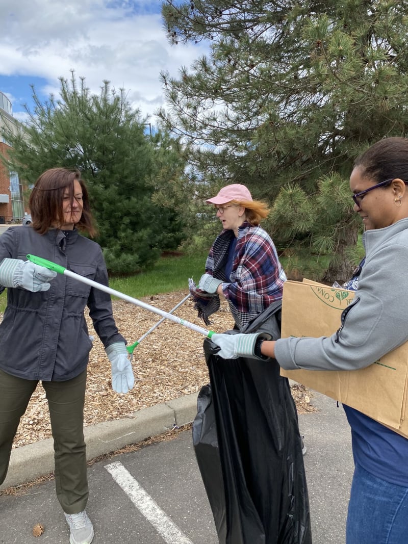 Three people wearing gloves use grabber tools to pick up litter outdoors near trees and a parking lot, holding a large black trash bag and a cardboard box.