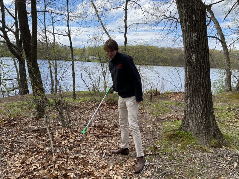 Person using a green and white metal detector outdoors on a leafy forest floor near a lake, wearing beige pants, dark jacket, and brown shoes.