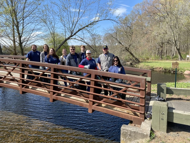 Group of people wearing blue shirts and gloves standing on a brown metal pedestrian bridge over a calm river, surrounded by trees and clear sky.