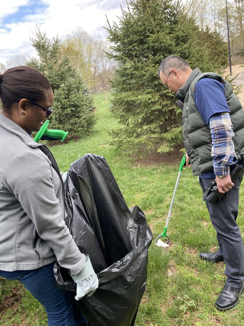 Two people outdoors using green-handled litter pickers to collect trash into a large black plastic garbage bag, surrounded by pine trees and grass.