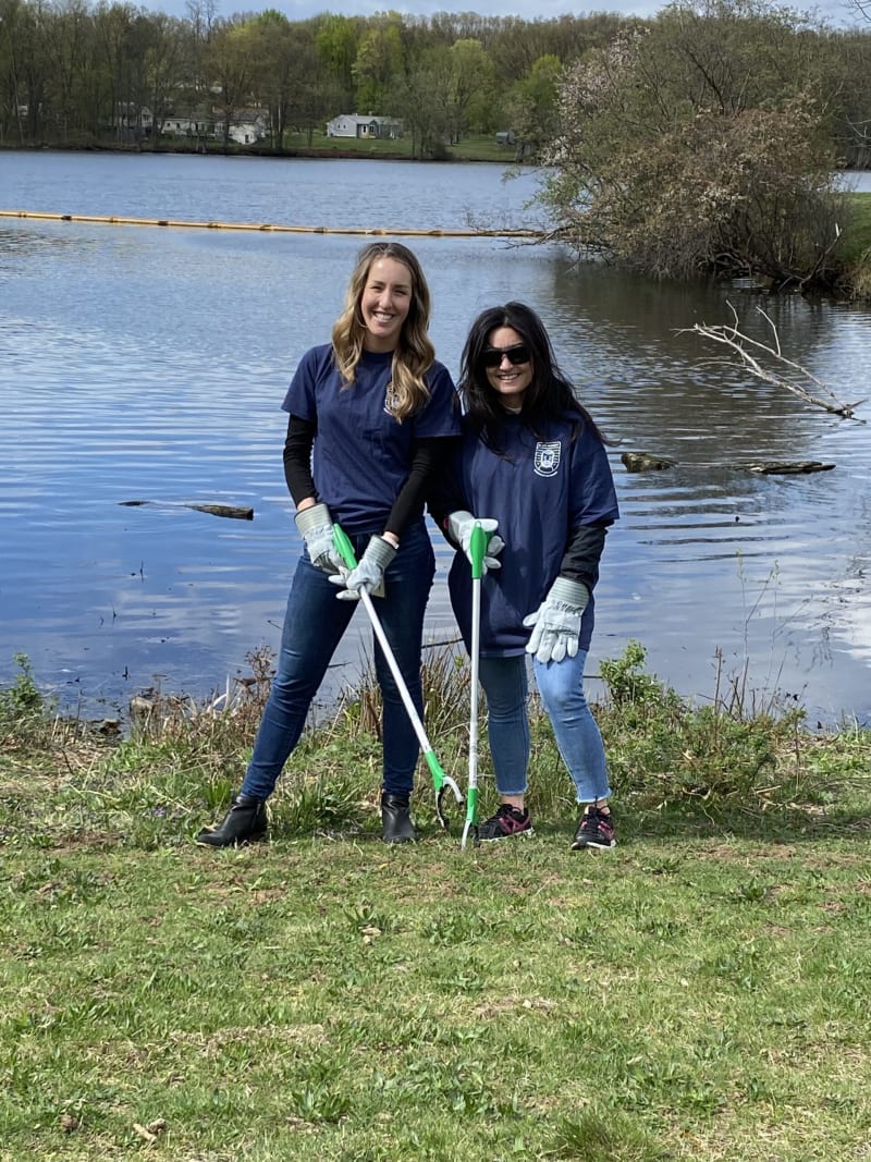 Two women wearing navy blue shirts, gloves, and jeans stand on grass by a lake holding green and white litter pickers, participating in outdoor cleanup.