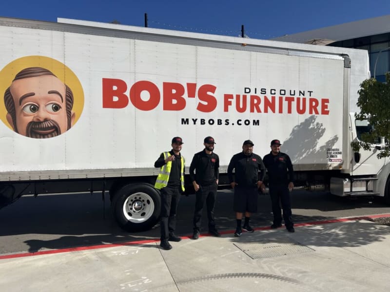 Four workers stand in front of a white delivery truck with large red text reading "BOB'S DISCOUNT FURNITURE" and the website "MYBOBS.COM" on the side, promoting furniture delivery services.
