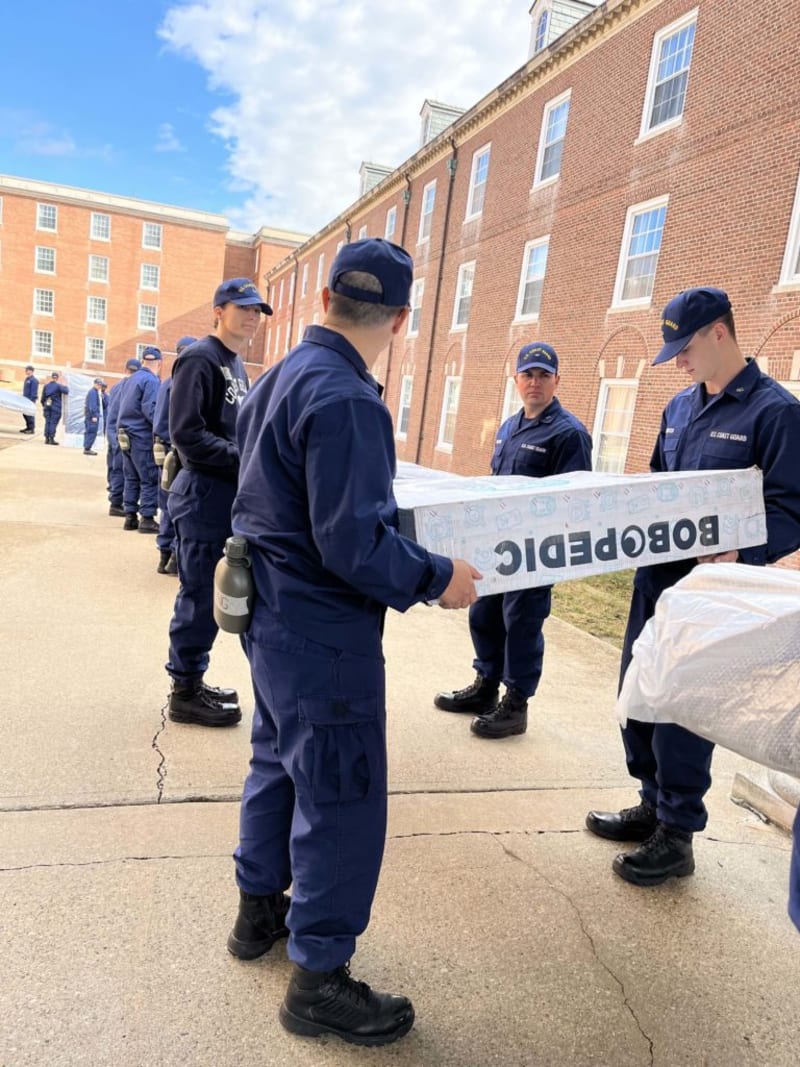 Uniformed personnel in navy blue Coast Guard attire are passing a large rectangular box labeled "BOBOPEDIC" outdoors near a brick building under a partly cloudy sky.
