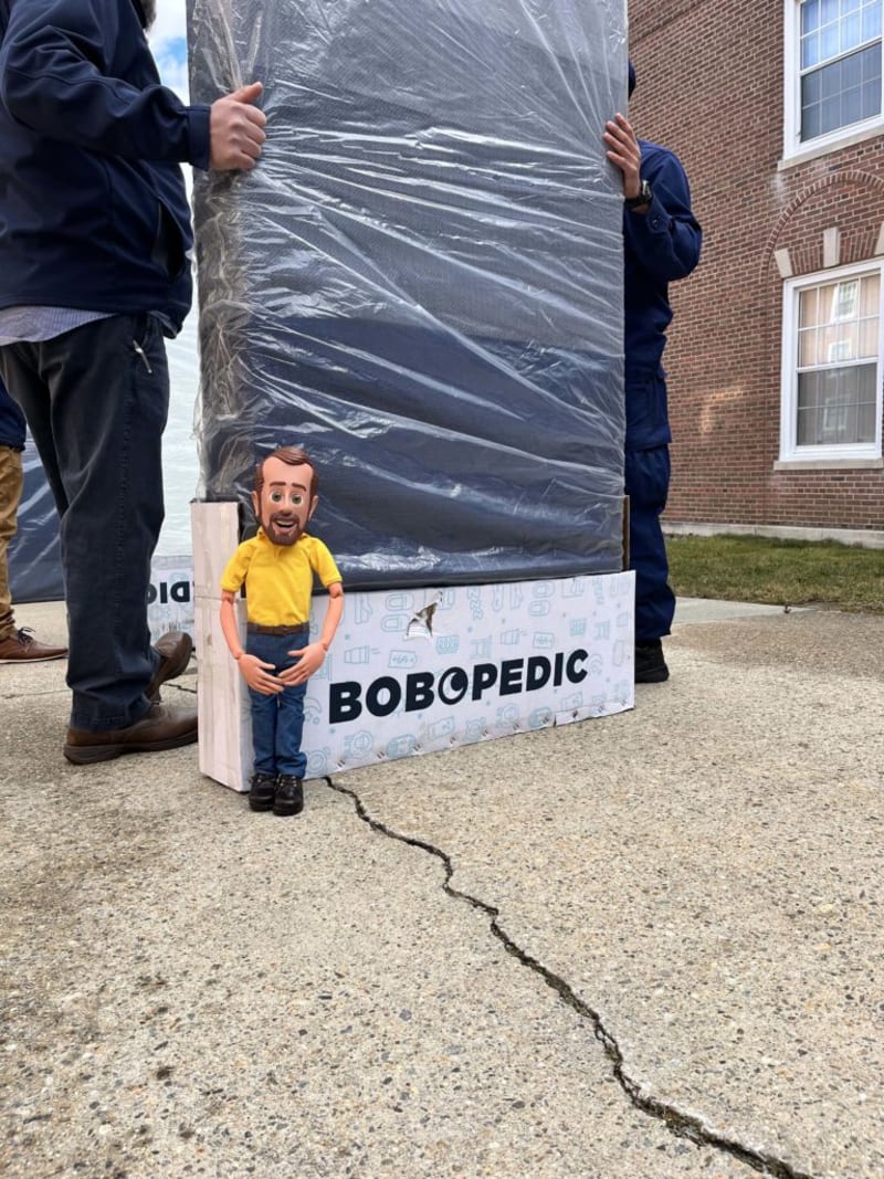Two people holding a large, plastic-wrapped mattress with a white box labeled "BOBOPEDIC" on a concrete surface near a brick building.