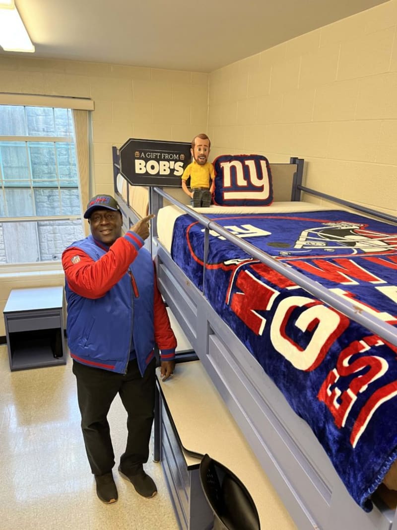 Man wearing a blue and red jacket and New York Giants cap points to a lofted bed with New York Giants bedding, pillow, and a sign reading "A GIFT FROM BOB'S."