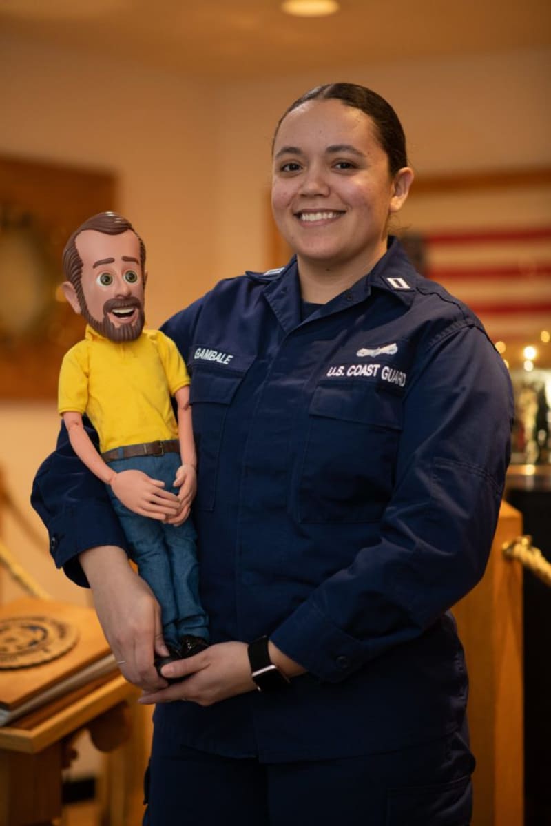 U.S. Coast Guard member in navy blue uniform holding a ventriloquist dummy wearing a yellow shirt and blue jeans indoors.