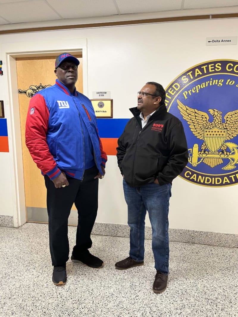 Two men standing indoors near a door labeled "DAYROOM E109 BOS'N Hole" and a large United States Coast Guard emblem with the text "Preparing to CANDIDATE." One man wears a blue and red New York Giants jacket and matching cap, black pants, and black shoes. The other wears a black jacket with red "BOB'S FURNITURE" logo, blue jeans, and brown shoes. The setting appears to be a formal or institutional environment.