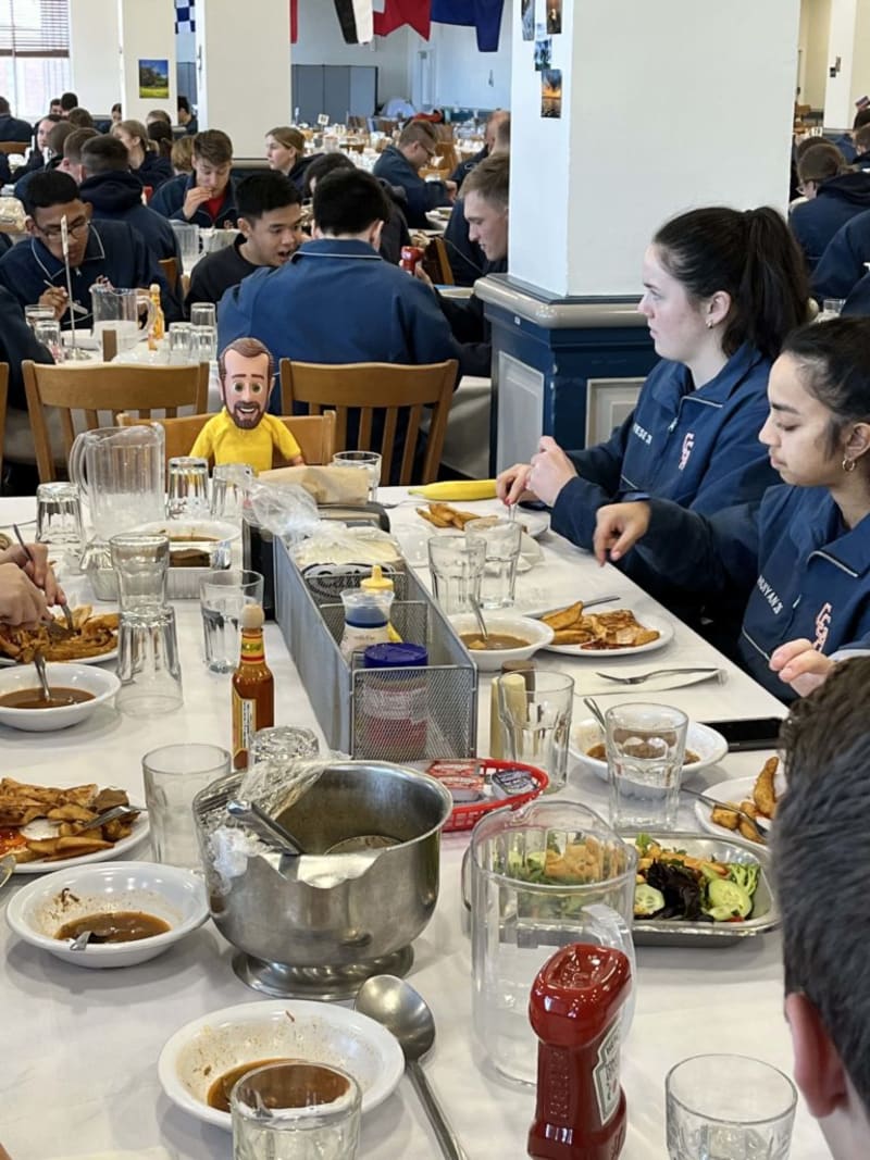 A busy cafeteria setting with people wearing navy blue uniforms seated at long tables, eating meals including fries, salad, and beverages, with condiments like Heinz ketchup and hot sauce on the white tablecloths.