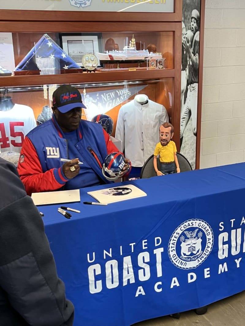 Man in New York Giants jacket signing a helmet at a United States Coast Guard Academy table with memorabilia and sports items in the background.