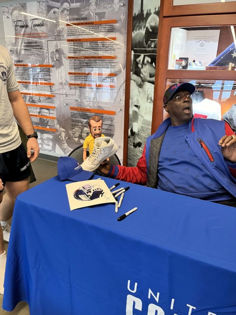 Man seated at a blue United States-themed tablecloth signing a white sneaker with black markers, wearing a blue and red jacket and cap, with a blue cap and a football helmet drawing on the table.