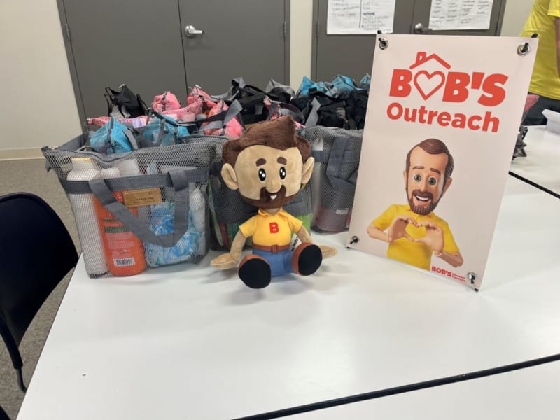 Bob's Outreach display with mesh bags filled with personal care items, a plush mascot wearing a yellow shirt with a red "B," and a sign featuring the text "BOB'S Outreach" in red with a heart-shaped house logo.