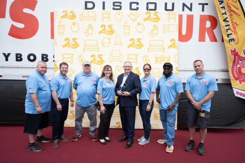 Group of eight people standing on a red carpet in front of a backdrop with yellow 25th anniversary icons and a large banner with "DISCOUNT" and "BOB'S" text, wearing light blue polo shirts with a red "25" logo, celebrating Bob's 25th anniversary event.