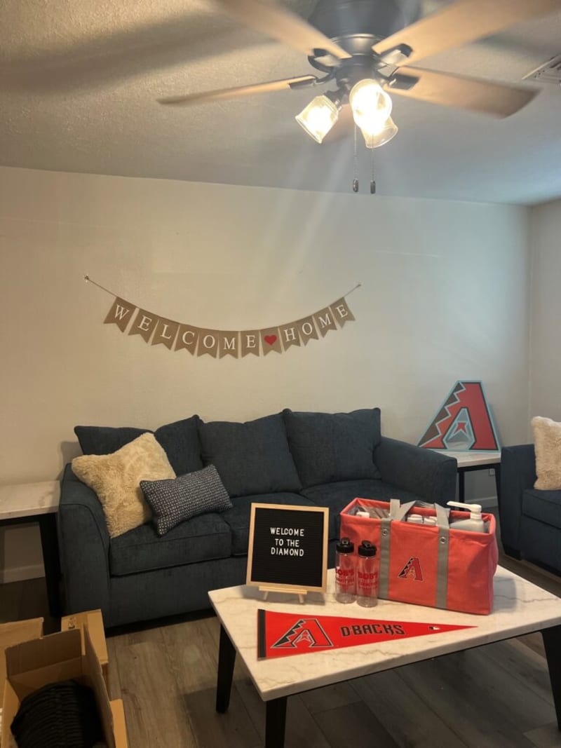 Dark blue fabric sofa with beige and patterned pillows under a "WELCOME ♥ HOME" banner, next to a white marble coffee table holding a red Arizona Diamondbacks bag, water bottles, and a sign reading "WELCOME TO THE DIAMOND."